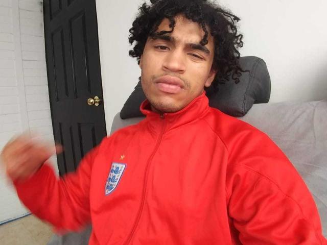Christopher Wood, um sujeito de cabelo cacheado, com pele marrom-clara e um bigode discreto, veste uma jaqueta vermelha com um emblema azul, piscando e levantando o punho direito..