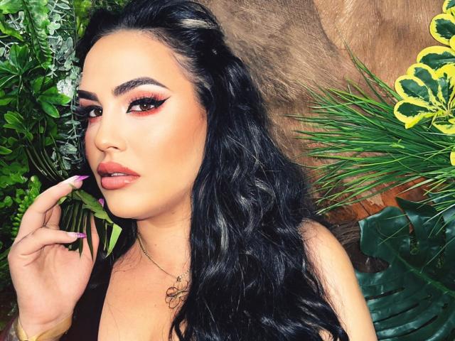 HottyNicole met lang, zwart, golvend haar, volle rode lippen en dramatische eyeliner, houdt groene bladeren vast, heeft roze nagels en een gouden ketting, en kijkt verleidelijk in de camera..