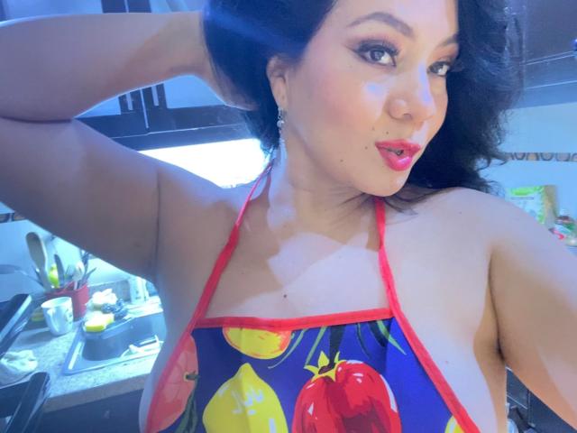 AlisGourmande with dark curly hair, red lipstick, and a colorful apron featuring fruit, showing her tits in a kitchen, hand in hair, blue light, earrings, and a cute smile.