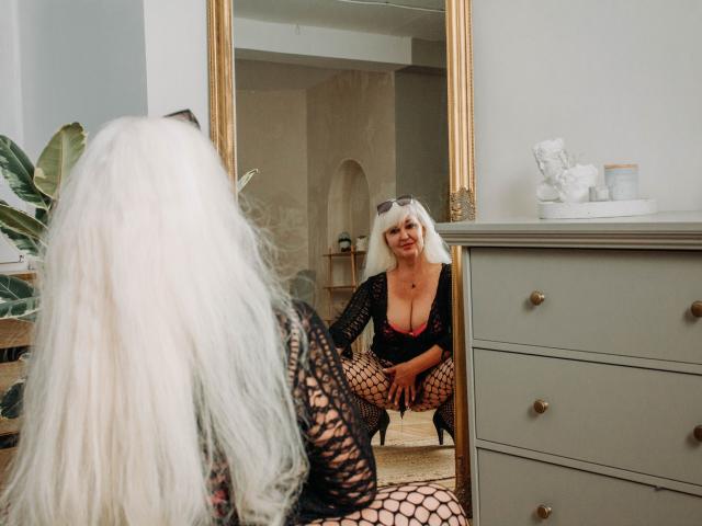 KamaliaSim with long white hair, black fishnet top, deep cleavage, and fishnet pants, crouching in front of a mirror, showing her tits and ass, in a room.