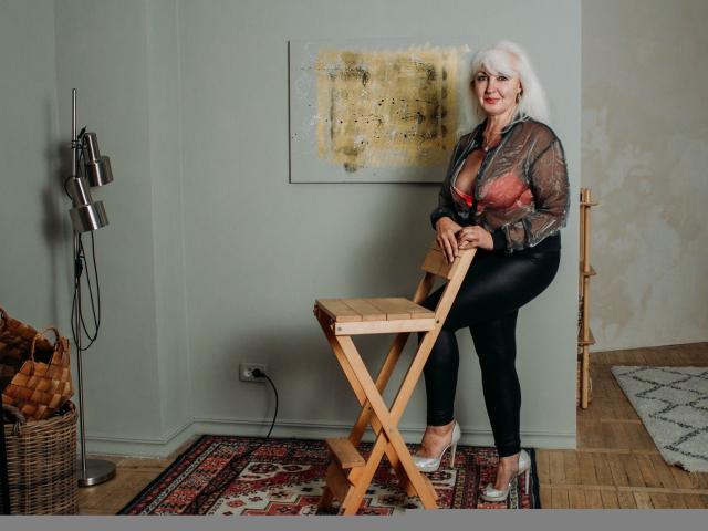 KamaliaSim, con cabello blanco, blusa roja, pantalones de cuero negro y tacones plateados, apoyada en un taburete de madera, mostrando sus grandes senos, de pie en una habitación con pared verde y alfombra..