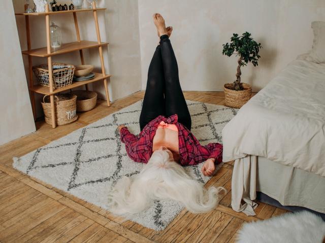 KamaliaSim, avec ses longs cheveux blancs, son legging noir et sa chemise à carreaux rouges, est allongée sur le dos, les jambes en l'air, exposant ses petits seins nus, avec des mamelons roses, sur un plancher en bois..
