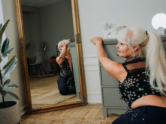KamaliaSim with white ponytail, black star-patterned top, flexing arm in front of mirror, showing reflection, wearing dark jeans, kneeling, wooden floor, potted plant on left.