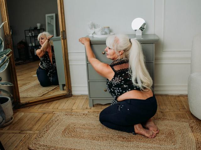 KamaliaSim with white hair in a ponytail, wearing a black floral top and dark jeans, kneels on a rug, touching a gray dresser, reflected in a mirror.
