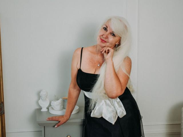 KamaliaSim with long white hair, black dress, white bow, leaning on gray table, smiling, hand on chin, small bust, white bust and vase, light skin, red lipstick.