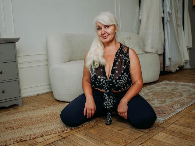 KamaliaSim, met wit haar, draagt een zwarte top met bloemenprint, die ze vooraan vastbindt, en donkere broek. Ze knielt op een houten vloer, glimlachend, waarbij haar kleine borsten en licht gebogen billen zichtbaar zijn..