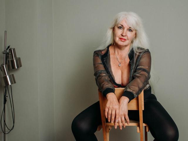 KamaliaSim with long white hair, sheer black top, exposing her big tits, black pants, sitting on a chair, hands on wooden seat, gray wall, metal wall art.