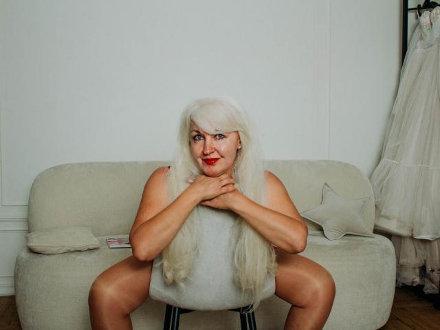 KamaliaSim with long white wig, red lipstick, sitting on a chair, legs spread, hands on chest, gray sleeveless top, white couch, pillow, and dress on a stand in background.