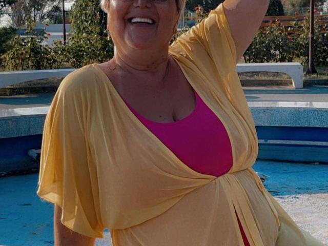 QueenPammy, a chubby older woman with short gray hair, wearing a yellow dress over a pink bra, smiling with her right arm raised. Her tits are visible under the dress.
