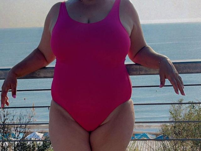 QueenPammy, with big boobs, wide hips, and a thick ass, wearing a pink one-piece swimsuit, leaning on a metal railing, ocean in the background, sunlight on her skin.