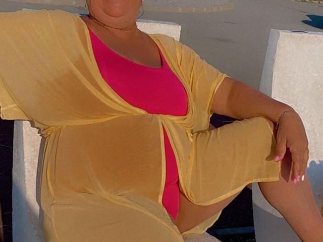 QueenPammy with tan skin, wearing a sheer yellow robe over a pink bikini, showing her ass and pussy, sitting with one leg up, nail polish visible.
