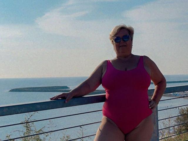 QueenPammy, a chubby woman with short blonde hair, wearing a pink one-piece swimsuit and sunglasses, leans on a railing with her hand on her hip. Her ass is visible.