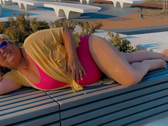 QueenPammy lying on a blue bench, wearing a yellow shirt and pink bikini, showing her tits and ass, with sunglasses on, in a sunlit outdoor setting.