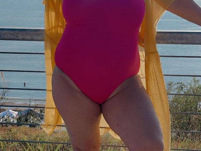 QueenPammy's pink one-piece swimsuit, yellow sheer top, and tan lines, showing her ass and thighs, standing by a metal railing with a beach and ocean in the background.
