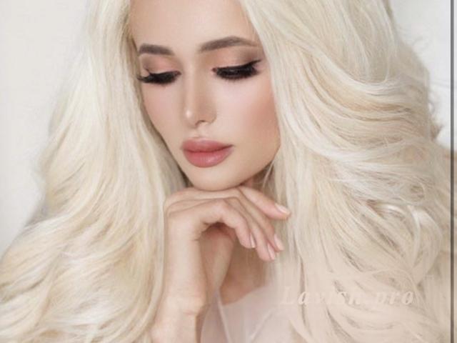 VikkiLike ha lunghi capelli biondi ondulati, rossetto rosa e guarda in basso con la mano vicino al mento. Ha l'eyeliner scuro e le unghie naturali..