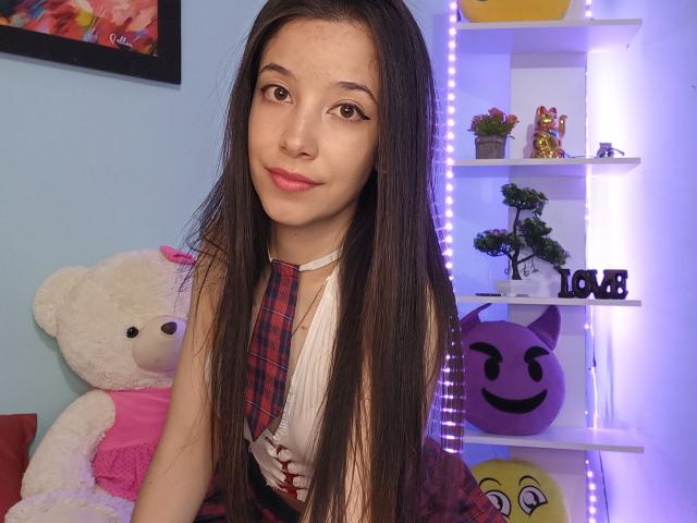 ManuMejia met lang bruin haar, roze lippenstift, een geruit das en een mouwloos shirt, lachend, een witte teddybeer links, een paars plankje met emoji's en een plant rechts..