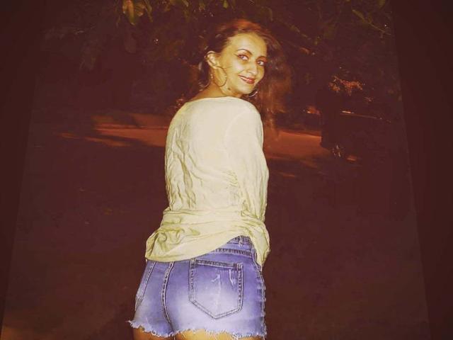 MelaniaTom, con capelli castani ondulati, sorridente, indossa una camicia gialla a maniche lunghe e pantaloncini di jeans blu sbiaditi, mostrando il sedere. Notte, sfondo scuro..