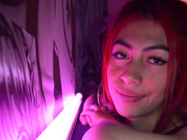 Dalyla, con i capelli rossi, orecchini a cerchio e un sorriso carino, mostra leggermente il seno illuminato da una luce rosa al neon. I suoi occhi sono socchiusi, con un'aria sensuale..