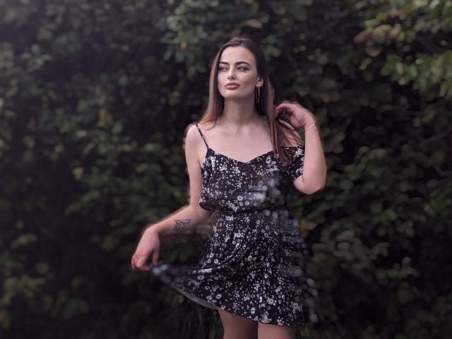 MilanaPrice with long brown hair, wearing a black floral dress, stands outdoors, holding her dress, showing a tattoo on her right arm, with a serious expression.