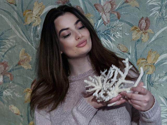 MilanaPrice with long brown hair, wearing a pinkish knit sweater, holding a white coral-like branch, looking down with a slight smile, soft pink lipstick, subtle makeup.