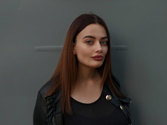 MilanaPrice, met bruin haar, een zwarte leren jas en een zwarte top, laat haar schattige gezicht en kleine neus zien, en staat daar met een subtiele glimlach. Haar borsten zijn klein..