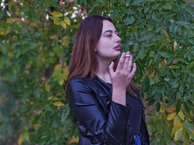 MilanaPrice, com cabelo castanho, jaqueta de couro preta, segurando um cigarro, fundo com folhagem verde, olhando para o lado, lábios levemente entreabertos, casual, sexy, estilosa, close do corpo superior..