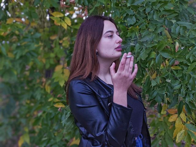 MilanaPrice met bruin haar, een leren jas, die een sigaret vasthoudt, met groene bladeren op de achtergrond, die naar de zijkant kijkt, met handen in de buurt van het gezicht, lichtjes open lippen, een serieuze uitdrukking..