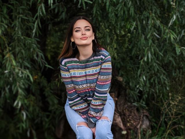 MilanaPrice with brown hair, wearing a colorful striped sweater and ripped jeans, sitting against greenery, smiling with eyes closed, wearing hoop earrings, and showing her tits slightly.