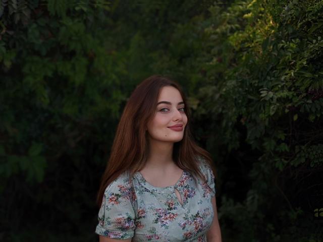 Milana, con lunghi capelli castani, un abito floreale, un sorriso carino e una piccola lentiggine sulla guancia destra, è in piedi di fronte a un fogliame verde scuro. Sta guardando a sinistra..