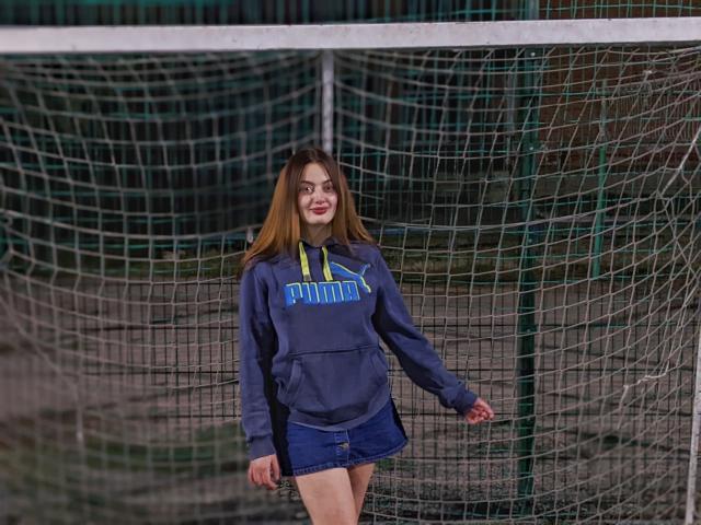 MilanaPrice stands in front of a soccer goal, wearing a blue Puma hoodie and denim skirt, smiling with long brown hair, her hands relaxed by her sides.