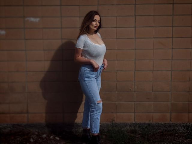 MilanaPrice with large boobs, wearing a tight white shirt, ripped light blue jeans, and black boots, standing against a brick wall, shadowed to the left.