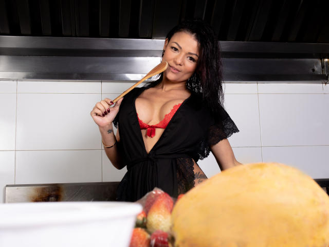 VeronicSaenz mit dunklem, nassem Haar, schwarzem Spitzenkleid, rotem BH, die einen Holzlöffel hält, im Vordergrund ein großes, gelbes Gebäck und Erdbeeren. Sie lächelt und zeigt ihre Brüste..