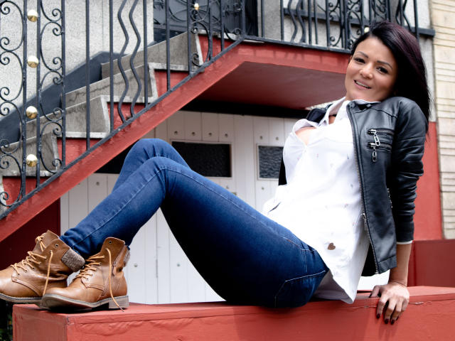 VeronicSaenz with tan boots, blue jeans, white shirt, black leather jacket, and dark hair, sitting on red steps, smiling, showing her ass, in front of a metal staircase.