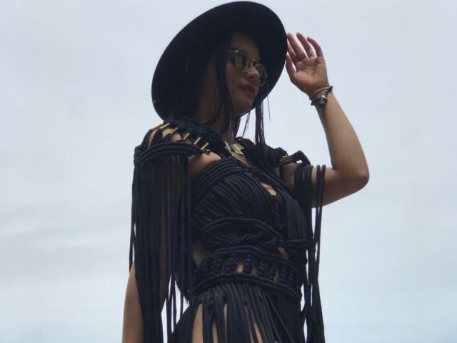 DahianMoretti, vêtue d'une robe noire à franges, laissant entrevoir ses seins à travers des découpes, portant un chapeau noir, des lunettes de soleil et un bracelet au poignet, levant la main vers un ciel nuageux..