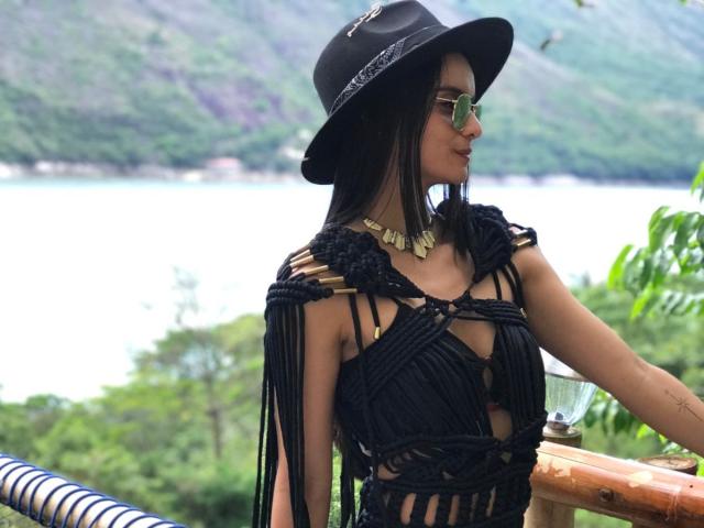 DahianMoretti, avec ses longs cheveux noirs, portant un chapeau noir, des lunettes de soleil vertes et un haut en crochet noir avec des franges, qui révèle ses petites seins et un collier en or..