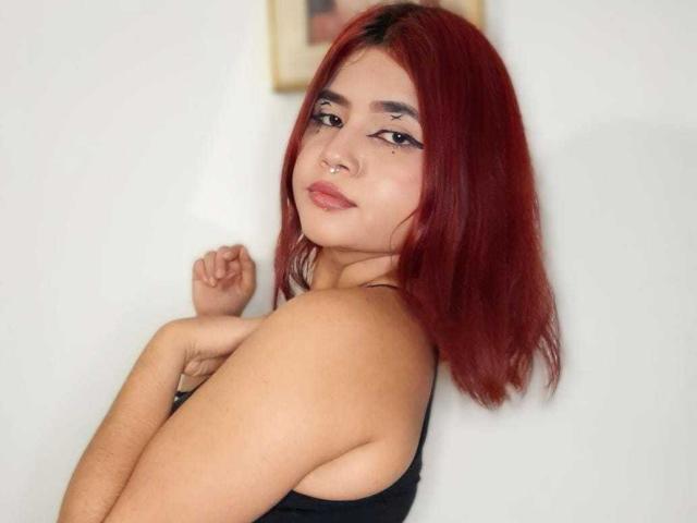 NicolRitz com cabelo ruivo, top preto, mostrando o bumbum, olhando por cima do ombro, sorriso discreto, olhos castanhos, maquiagem suave, parede branca, quadro na parede ao fundo..