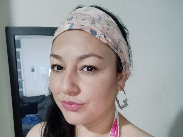 SakotoDika with a pink floral headband, black hair, brown eyes, pink lipstick, and silver earrings, smiling slightly, showing her nose and upper chest.