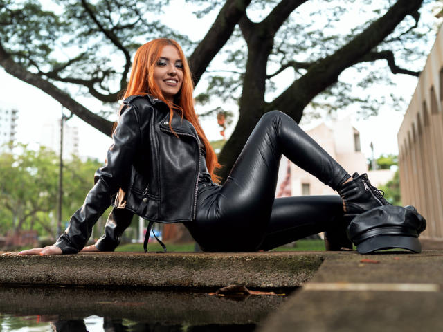 Maisy Warner, met lang rood haar, een zwarte leren jas en glanzende zwarte leren broek, zittend op een betonnen rand, lachend, met zwarte laarzen aan, met een boom op de achtergrond..