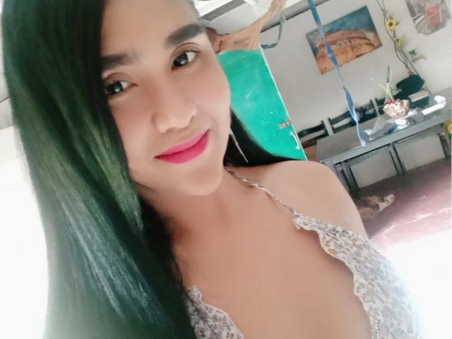 A MsHealthyCumTs tem cabelos pretos longos, batom vermelho e está vestindo uma blusa branca de renda com alças, que mostra seus pequenos seios, e está sorrindo para a câmera..