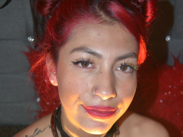 DalylaLove met rood haar, een zwart leren verstelbaar harnas, een rode bal-gag, kleine tatoeages, lachend, een rood, pluizig kussen, een rode muur en een grijze stoel op de achtergrond. Borsten zichtbaar..
