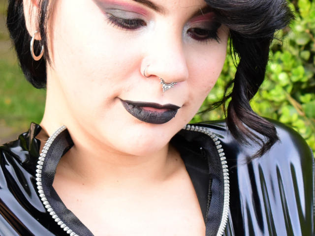 DalylaLove nue, avec des cheveux roux et des tatouages, à genoux, les poignets et les chevilles enchaînés, bouche attachée avec un entonnoir, les yeux fermés, la tête penchée en arrière, tenant une balle rouge dans la main, fond noir texturé..