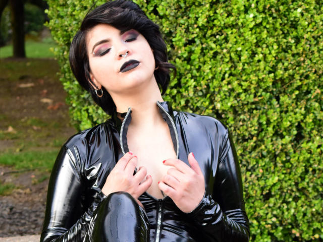 AndiChin, avec du rouge à lèvres noir, un anneau de nez et une veste en latex noir, portant des boucles d'oreilles cerceaux et un maquillage foncé, regardant vers le bas avec ses cheveux noirs bouclés. Lumière douce, fond vert..