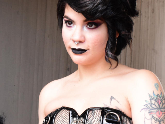 AndiChin, vêtue d'une combinaison en latex noir brillante, les mains sur ses seins, les yeux fermés, avec un rouge à lèvres noir, des cheveux noirs courts, des boucles d'oreilles pendantes, debout devant un buisson vert..