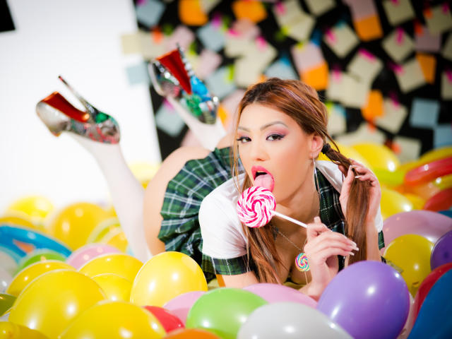 CataleyaLee mit langen braunen Haaren, einem grünen karierten Rock, einem weißen Hemd und High Heels, leckt an einem rosa gesprenkelten Lutscher, umgeben von bunten Ballons und trägt weiße Socken..