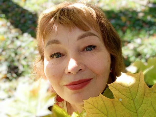 MilaMils, con i capelli castani corti, gli occhi azzurri e il rossetto rosso, sorride dolcemente mentre tiene una foglia gialla davanti al viso. I suoi seni sono leggermente visibili sotto la foglia..