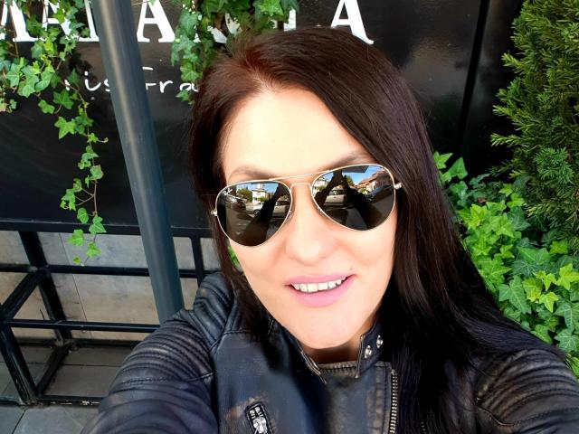 JoliDanielle with dark brown hair, wearing reflective aviator sunglasses and a black leather jacket, smiling at the camera, showing white teeth. Her pink lips are slightly parted.