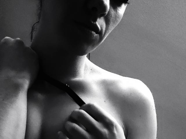 Le spalle scoperte di Esra, che tiene una tracolla, in una foto in bianco e nero, con un viso leggermente in ombra e un sorriso appena accennato, che mostra una parte superiore del corpo modesta, delicata e sensuale..