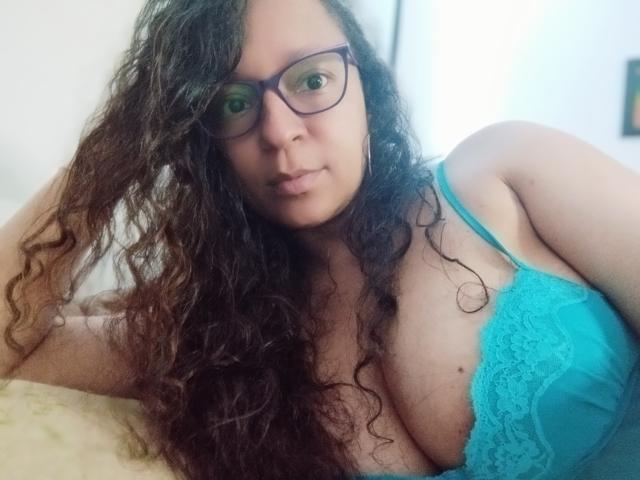 Angelonix with curly brown hair, glasses, and teal lace bra, showing her large tits, lying on a bed, looking into the camera with a slight pout.