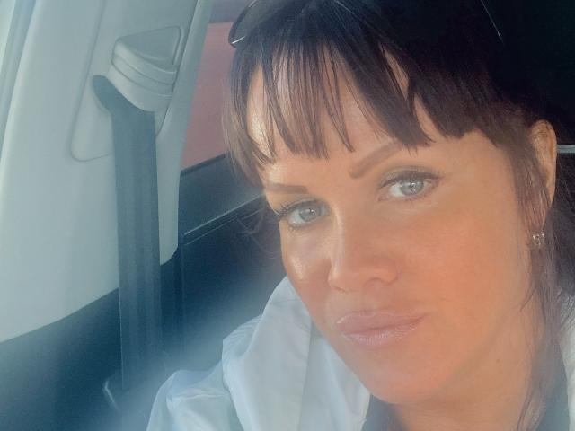 LolaleyS a les cheveux bruns avec une frange, des yeux verts, et une légère moue, elle porte une chemise blanche, est assise dans une voiture, a une expression neutre et un bronzage léger..