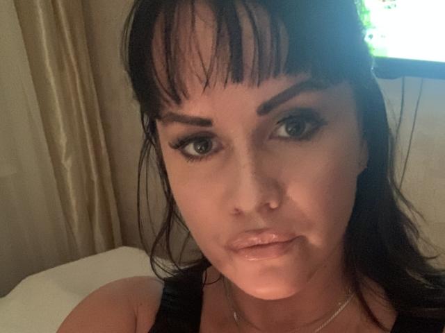 LolaleyS has dark brown hair with bangs, green eyes, and glossy pink lips. She's wearing black makeup and a silver necklace, looking directly at the camera with a slight smile.